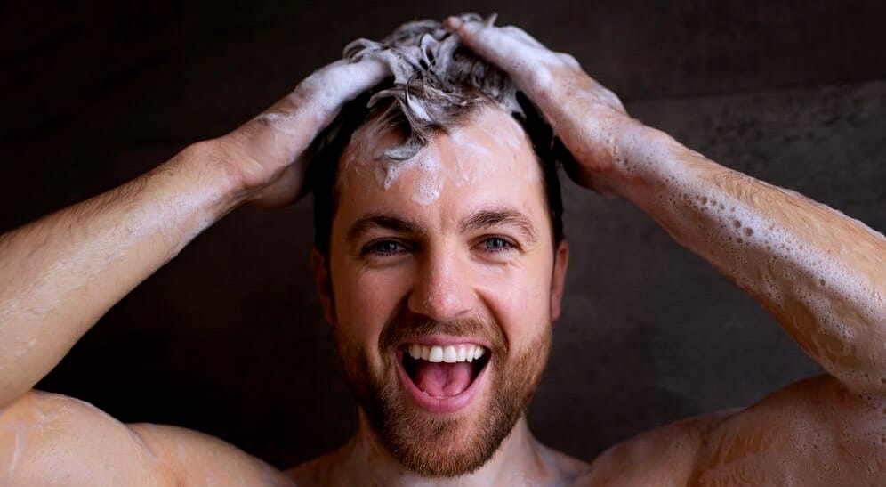 Uśmiechnięty mężczyzna pod prysznicem myjący głowę szamponem